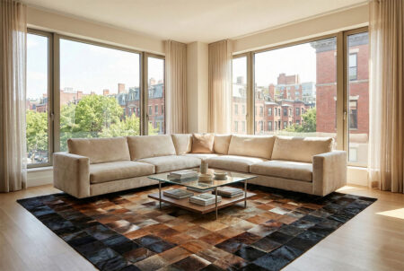 224-The-Gradient-Patchwork-Cowhide-Rug---Earth-Tones Modern luxury living room featuring a large patchwork cowhide rug with a black, brown, and beige gradient pattern, placed under a beige velvet sectional sofa near floor-to-ceiling windows