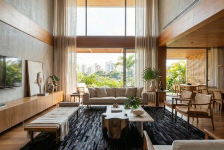 Black Striped Patchwork Cowhide Rug - Sun-Drenched Modern Living Room with a View 253 Open-plan living room with a super high ceiling and a massive window that looks out onto a green park. Sunlight streams in, illuminating a long, beige sofa, a couple of wooden armchairs, a daybed, and a cool, natural wood coffee table, all placed on a large, Black striped cowhide rug. There's a lot of wood on the ceiling and walls, and some plants, making it feel very warm and natural.