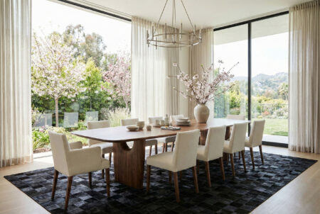 255-Black-Squares-Patchwork-Cowhide-Rug-in-Modern-Dining-Room Spacious modern dining room featuring a large black square patchwork cowhide rug placed under a solid walnut dining table and beige upholstered chairs. Big windows and blooming spring trees outside.