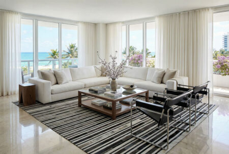 269-striped-patchwork-cowhide-rug-in-miami-beach-condo-marble-floors2 A sophisticated, sun-drenched living room with a Miami Beach aesthetic. The centerpiece is a large, horizontal striped patchwork cowhide rug in black, white, and grey. A white L-shaped cotton sofa and two black leather Wassily armchairs sit atop the rug, surrounding a wooden coffee table with a glass top. The room features shiny tiled flooring and a neutral, airy color palette.