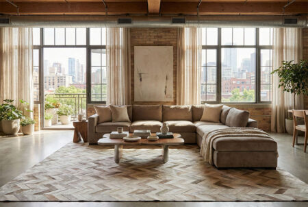 307-Modern-Industrial-Loft-Living-Room-with-Chevron-Cowhide-Rug A spacious, modern industrial loft living room featuring a large taupe and beige chevron patchwork cowhide rug. A beige velvet sectional sofa sits on the rug across from a modern wooden coffee table. The room features exposed brick walls, tall windows with sheer curtains, and a bright city skyline view.