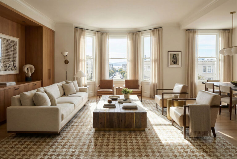 A spacious, luxury living room in San Francisco featuring a large, beige and white patchwork cowhide rug with a houndstooth pattern. Natural sunlight streams through tall bay windows with sheer curtains, highlighting the hair-on-hide texture of the rug. The space is styled with a long cream sofa, a blocky wooden coffee table, and leather accent chairs, creating a warm, neutral open-plan atmosphere