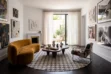 A brightly lit, luxurious living room featuring a round patchwork cowhide rug in a grey and white houndstooth pattern centered on a dark wooden floor. To the left is an organic, semi-circle sofa in ochre velvet, facing a dark wood, kidney-shaped coffee table. On the right sits an Italian Postmodern armchair with a geometric upholstery pattern. The white walls are decorated with modern art and neutral relief sculptures, alongside a marble fireplace. Floor-to-ceiling sheer curtains frame an open glass door leading to a sunny outdoor patio.