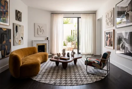 Ochre-velvet-and-houndstooth-cowhide-area-rug A brightly lit, luxurious living room featuring a round patchwork cowhide rug in a grey and white houndstooth pattern centered on a dark wooden floor. To the left is an organic, semi-circle sofa in ochre velvet, facing a dark wood, kidney-shaped coffee table. On the right sits an Italian Postmodern armchair with a geometric upholstery pattern. The white walls are decorated with modern art and neutral relief sculptures, alongside a marble fireplace. Floor-to-ceiling sheer curtains frame an open glass door leading to a sunny outdoor patio.