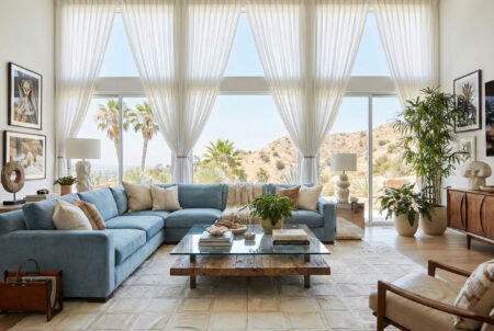 White-Mona-Patchwork-Cowhide-Rug-Blue-Velvet-Sofa-LA-Style Large White Mona patchwork cowhide rug in a luxury eclectic living room, placed under a powder blue velvet sectional sofa and glass coffee table near floor-to-ceiling windows.