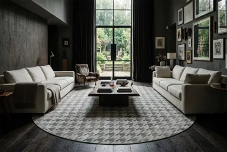 houndstooth-gray-and-white-cowhide-rug-in-eclectic-moody-living-room A luxurious, moody living room featuring a large, round patchwork cowhide rug with a grey and beige houndstooth pattern. The space includes dark-toned walls, high ceilings, dark wooden floors, two light linen sofas facing each other, a rustic wood coffee table, and an eclectic armchair. A curated gallery wall of art hangs on the right, and large, tall glass doors open to a lush green garden in the background, letting in natural light.