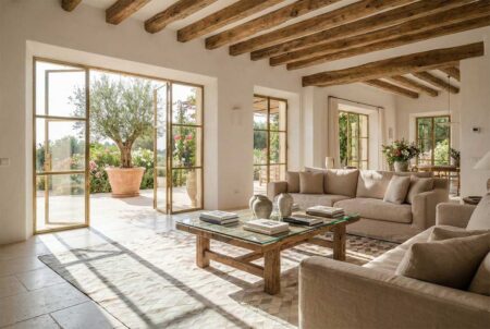310-Lime-ombre-cowhide-rug-in-franch-mallorcan-living-room Architectural shot looking into a modern living room, framed by gold-toned metal glass doors. The interior features high ceilings with massive rustic timber beams, white plaster walls, and a neutral color palette. A geometric cowhide rug anchors the center of the room, sitting beneath a rustic wooden coffee table and linen sofas, illuminated by bright natural sunlight.
