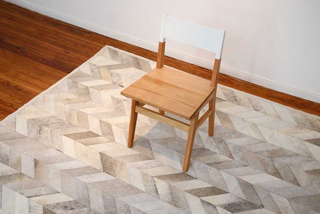 Corner view of a gray cowhide patchwork rug in a chevron design with a wooden chair on