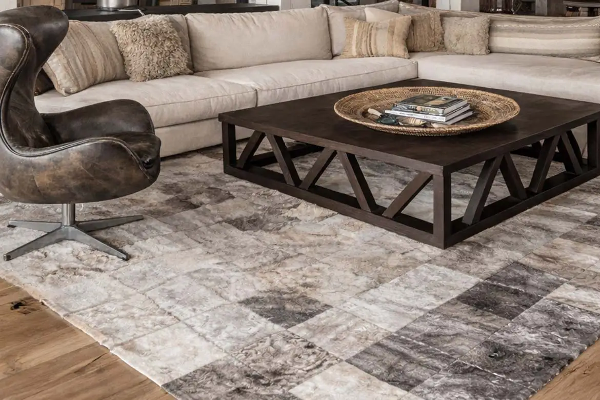 A refined western living room with exposed wood beams, neutral textures and layered natural materials. A patchwork cowhide rug in gray squares anchors the whole room. Modern western decor focused on structure, warmth and architectural balance. A gray squares patchwork cowhide rug rests under a heavy wooden table, anchoring the room.