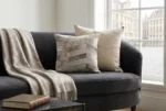 Elegant living room corner featuring a dark gray velvet curved sofa styled with a soft beige throw blanket, a neutral textured cushion, and a prominent gray and beige chevron cowhide patchwork pillow. Sunlight filters through open sheer curtains by a large window, with framed abstract art on the left walll.