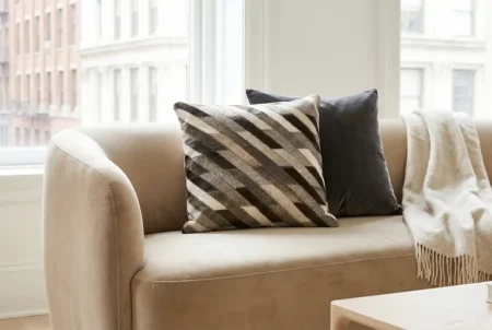 01 Stripes cushion cowhide cover in hotel lounge A close-up photograph of a contemporary beige velvet sofa positioned near two large windows overlooking a city cityscape. Arranged on the sofa are two decorative throw pillows: one in the front with a dynamic, diagonal chevron pattern in natural hide (black, grey, and cream fur), and a solid, charcoal-grey velvet pillow behind it. A light-colored, fringed throw blanket is draped casually over the armrest. A partial view of a light-wood coffee table is in the foreground. Natural light fills the room.