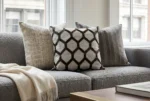 An elegant living room setting featuring a textured gray boucle sofa next to a bright window. The sofa is styled with three decorative cushions, centering on a geometric hexagon-patterned pillow made of brown cowhide and woven fabric. A soft, fringed cream throw blanket rests on the seat, with a partial view of a brass coffee table in the foreground.