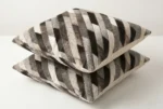 Pair of square, patchwork leather cushions made from distinct, diagonal stripes of hair-on hide, creating a striped pattern. The hide segments feature varying natural colors, including dark taupe, medium grey, light grey, and cream/white fur textures, sewn together in precise diagonal bands.