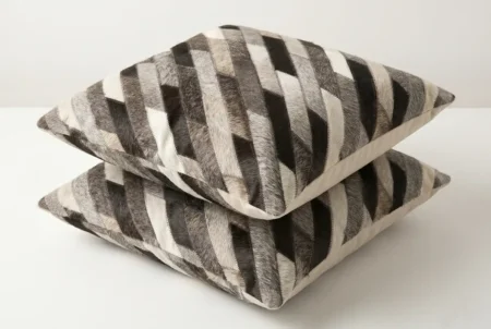 02 Stripes cowhide cushions in cream taupe gray Pair of square, patchwork leather cushions made from distinct, diagonal stripes of hair-on hide, creating a striped pattern. The hide segments feature varying natural colors, including dark taupe, medium grey, light grey, and cream/white fur textures, sewn together in precise diagonal bands.