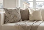 An interior photograph captures a corner of a sophisticated cream sofa. Three decorative pillows are arranged: the leftmost cushion is square and features a textured, geometric pattern of hide and fabric in cream, beige, and grey; the central pillow is a plain, textured neutral taupe; and the rightmost is a plain, textured off-white. A fringed cashmere throw blanket in a matching neutral shade is draped across the sofa seat. Behind the sofa, a large multi-pane window frames a classic view of Parisian Haussmann buildings and roofs under a soft, diffused light. The overall palette is composed of natural, elegant neutrals.