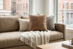 A modern tan linen sofa styled with a mocha brown leather patchwork cushion, neutral woven pillows, and a fringed throw blanket next to a bright window.